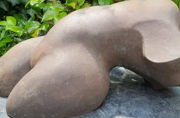 Kim Kroes- Ode aan de vrouw. Abstract bronzen sculptuur met ronde vormen, omringd door groene bladeren.